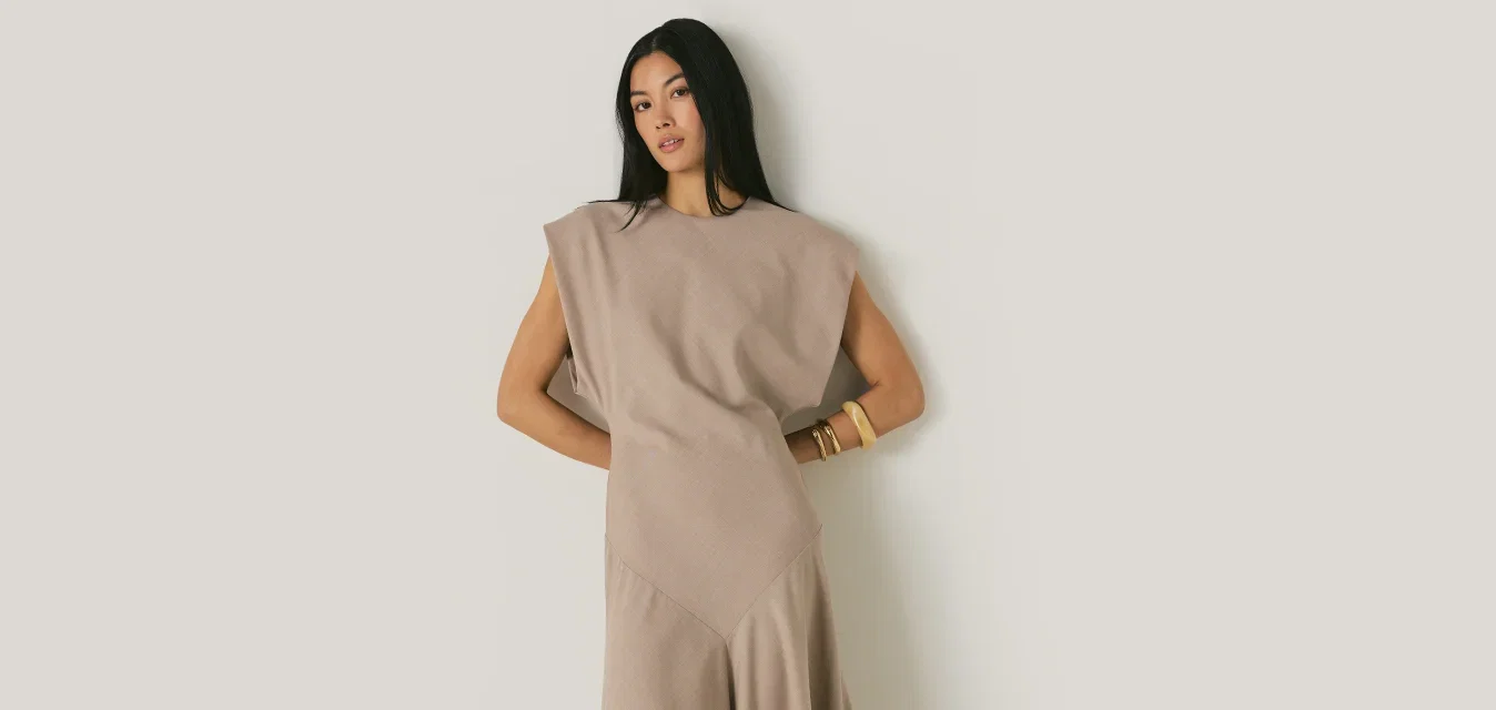 Femme aux longs cheveux noirs portant une robe midi beige sans manches avec des épaules rembourrées et des mocassins noirs pointus, debout contre un mur blanc.