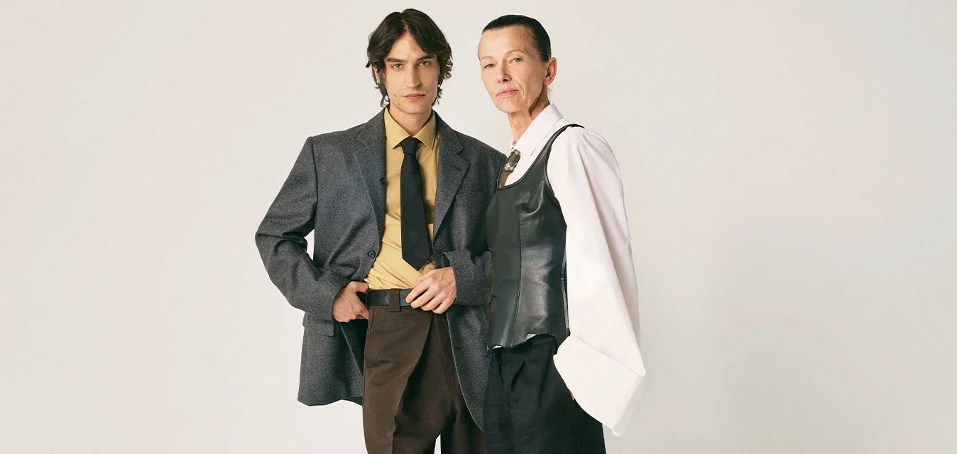 Deux adultes se tiennent devant un fond uni, vêtus d'une tenue formelle élégante : l'un en blazer gris avec une cravate, l'autre en blouse blanche avec un gilet noir.