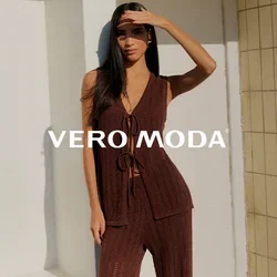 Femme en haut sans manches marron tricoté et pantalon, debout pieds nus sur une terrasse ensoleillée, se protégeant les yeux de la main contre un ciel bleu clair.