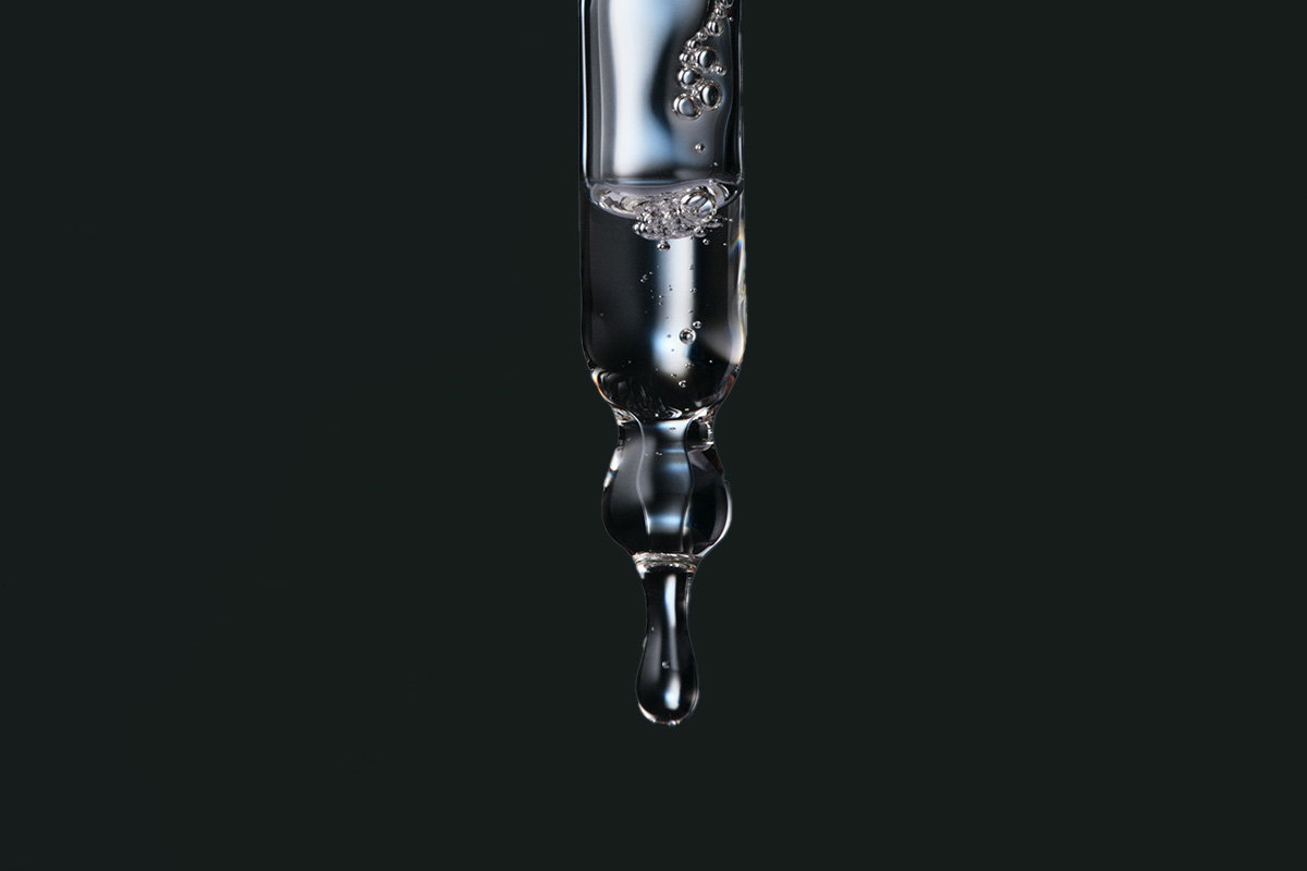 Close-up van een glazen pipet gevuld met heldere vloeistof tegen een donkergrijze achtergrond, klaar om een druppel te laten vallen.