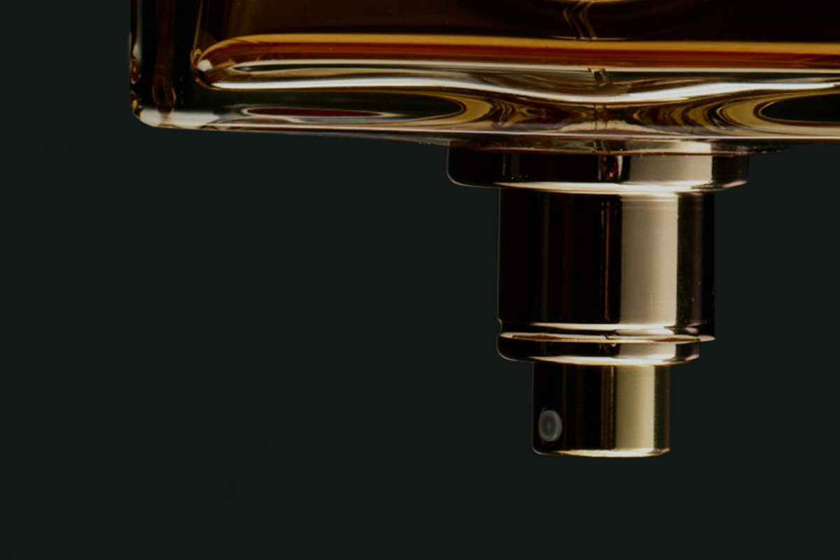 Close-up van de gouden verstuiver van een parfumflacon tegen een donkere achtergrond, wat een vleugje luxe en verfijning suggereert.