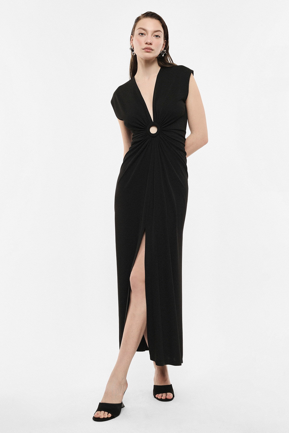 Woman modelling black sleeveless evening dress with central ring twist, deep V-neck and thigh-high slit, black heeled mules