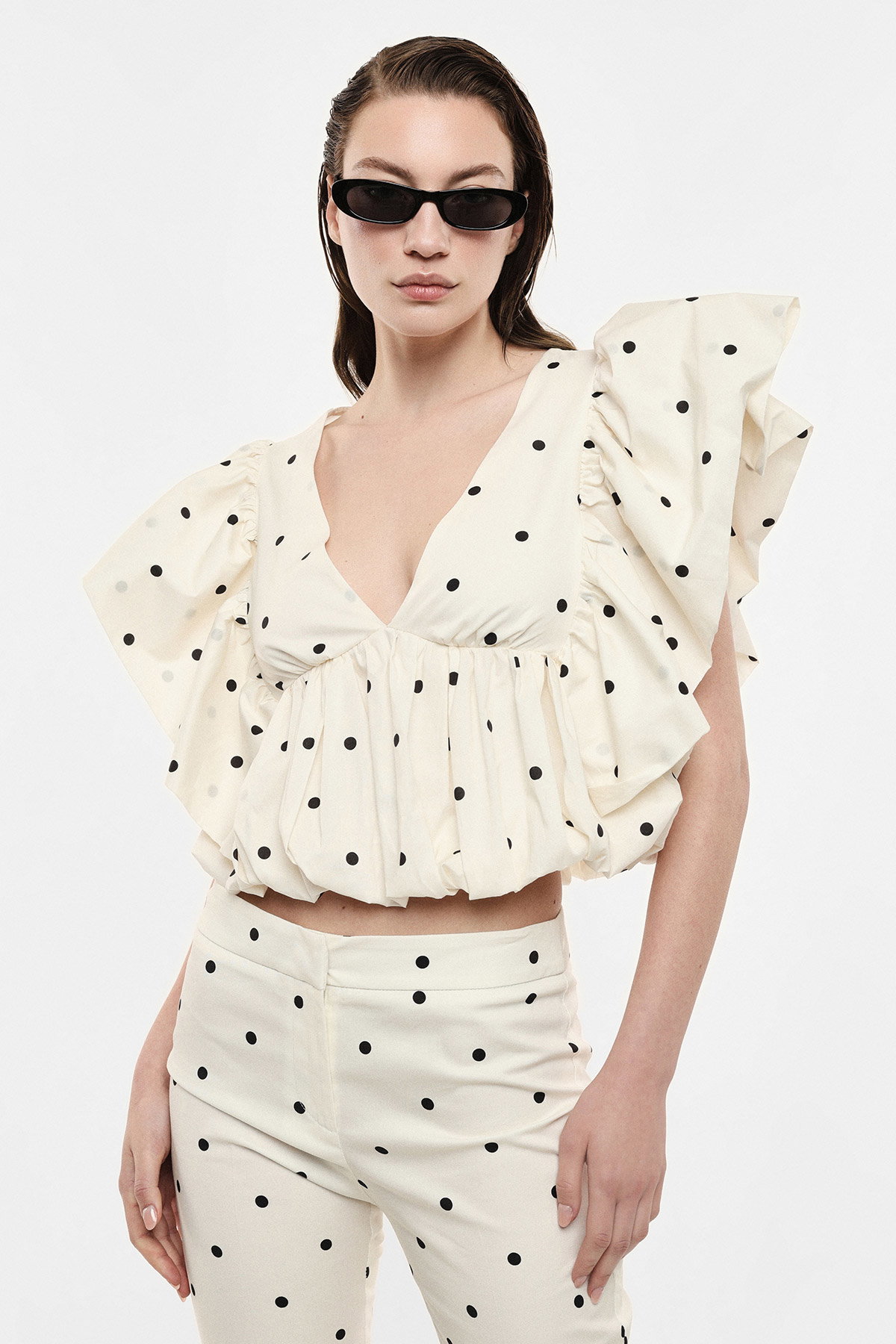 Woman wearing cream polka-dot ruffled crop top and matching high-waisted trousers with slim black sunglasses on a plain background
