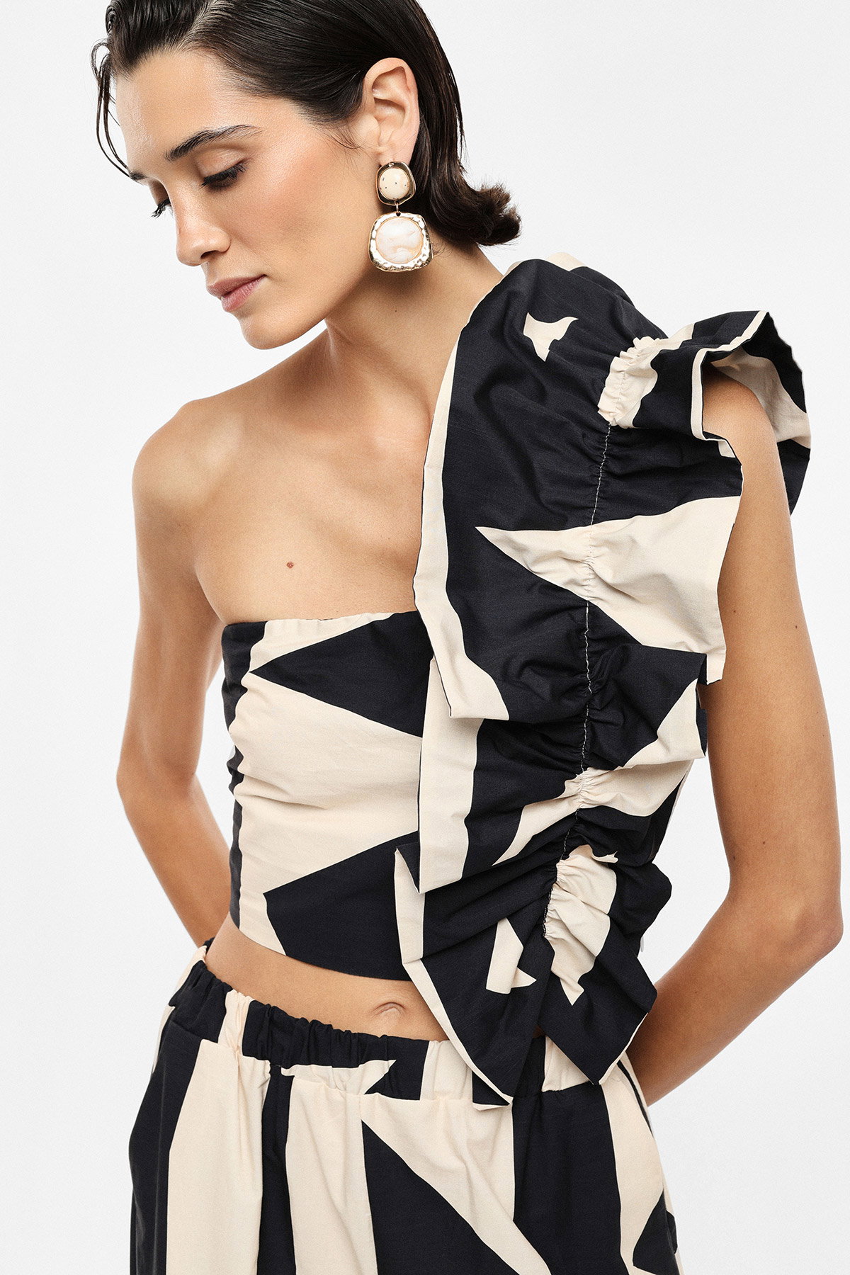 Model wearing black and cream geometric one-shoulder ruffled crop top and matching skirt with statement earrings, looking down