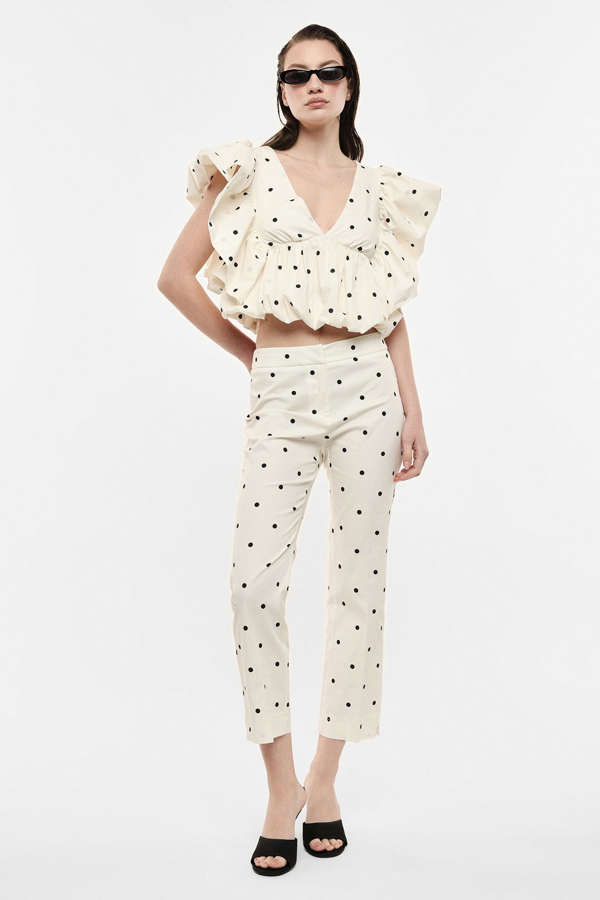 Model wearing cream polka-dot co-ord: ruffled V-neck crop top and matching high-waist trousers, black sunglasses and heels