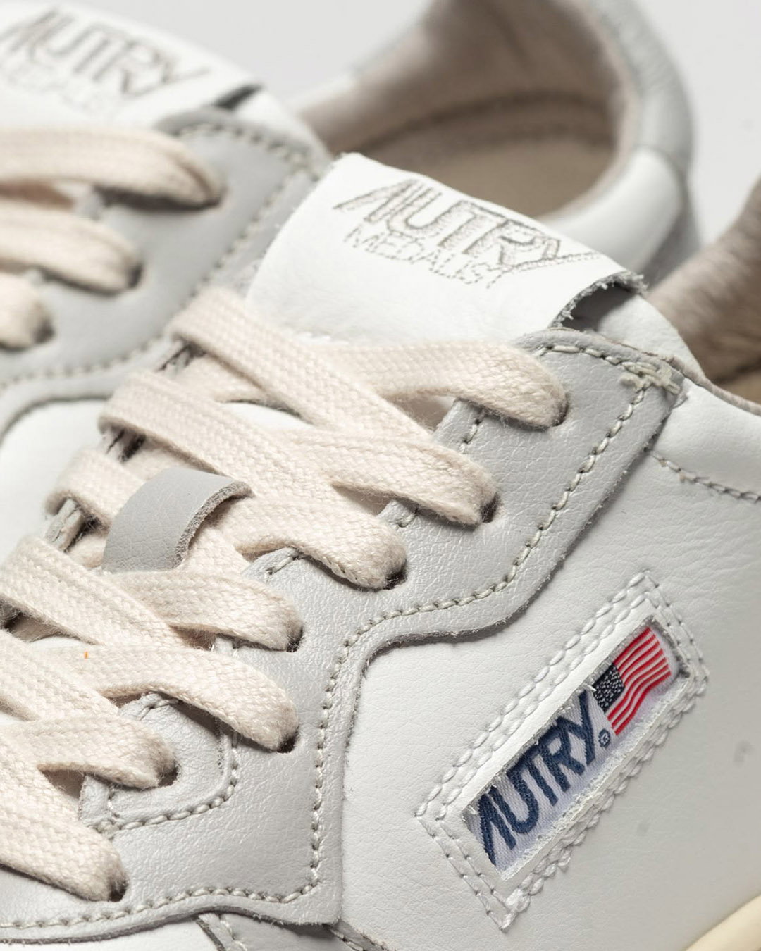 Close-up of white leather Autry trainers showing cream laces, stitched side logo with American flag and padded collar