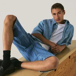 Joven recostado en una caja de gimnasia de madera, vistiendo pantalones cortos de mezclilla, camisa de mezclilla, camiseta blanca, calcetines negros y mocasines negros.