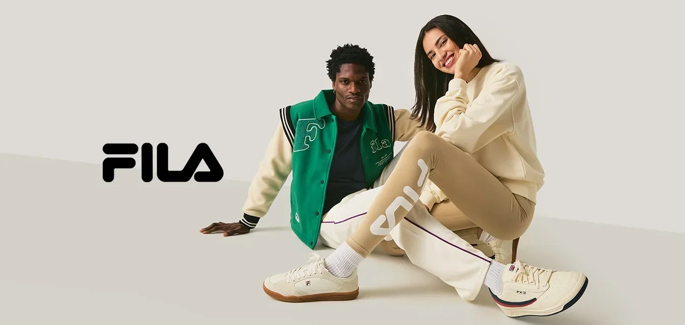 Dos personas sentadas en el suelo, una con una chaqueta varsity verde y blanca, la otra con mallas beige y un suéter crema, sonriendo.