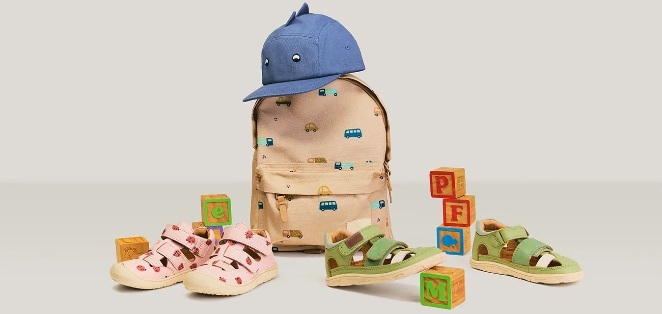 Mochila beige para niños con estampados de vehículos, gorra de béisbol azul encima, sandalias rosas y verdes, y bloques de alfabeto de madera dispuestos cerca.