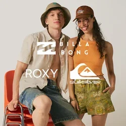 Joven con pantalones cortos y sombrero tipo pescador sentado sobre sillas rojas apiladas junto a una mujer sonriente con top naranja y pantalones cortos con estampado tropical.