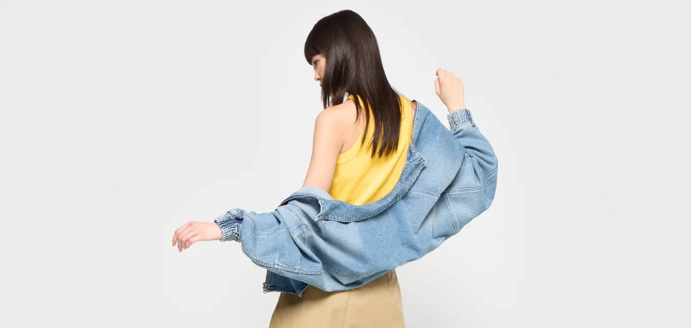 Giacca di denim, azzurro chiaro, vestibilità oversize; top giallo; gonna beige; mule con tacco marroni; in piedi con la giacca appoggiata sulle braccia, di spalle.