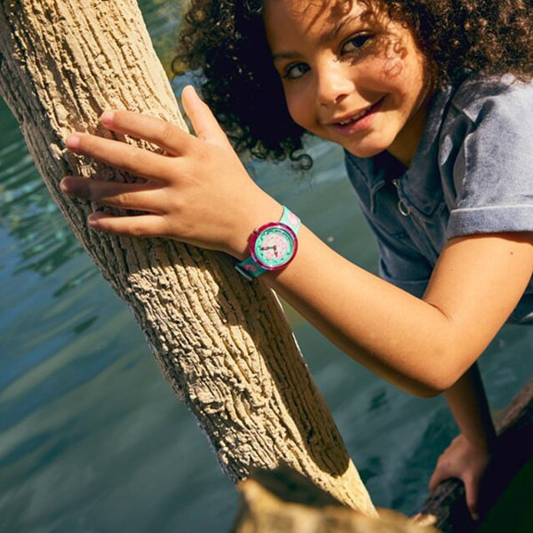 Kind am Wasser, hält sich mit der Hand an einem Baum fest. Am Handgelenk trägt sie eine Flik Flak Armbanduhr