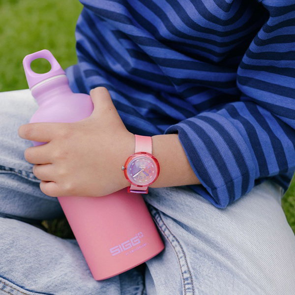 Kind hält eine Getränkeflasche in der Hand. Am Handgelenk tärgt sie eine Flik Flak Armbanduhr. Uhr und Flasche sind Ton in Ton rosa und lila