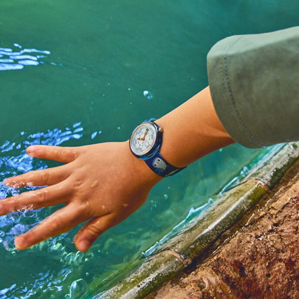 Die Kinderhand greift in ein Gewässer. Das Kind trägt am Handgelenk eine Flik Flak Armbanduhr in den Farben blau und grün