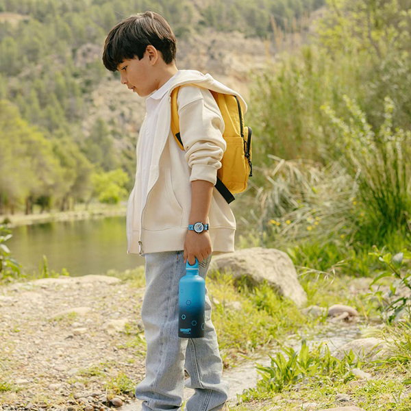 Kind mit Rucksack und Trinkflasche trägt eine Flik Flak Uhr am Handgelenk und läuft an einem Fluss entlang