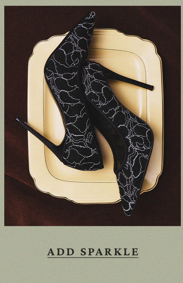 Black pointed-toe pumps with a white lace-like pattern resting on a cream scalloped tray against a dark brown velvet background. Text: ADD SPARKLE.