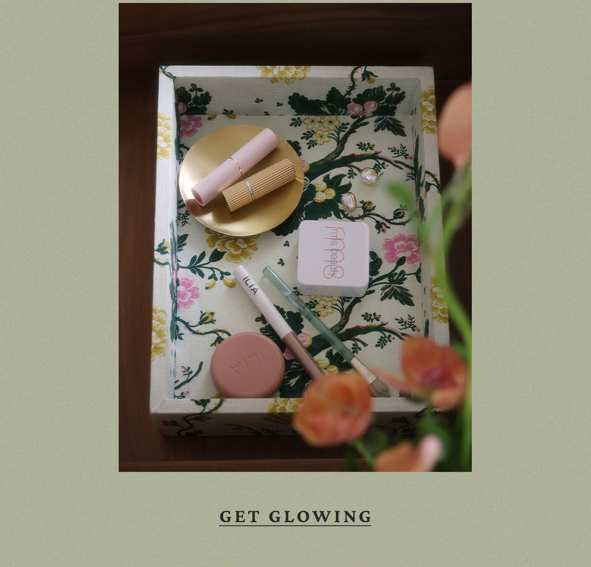 An overhead view of makeup products, including ILIA and RMS Beauty, displayed in a floral-lined tray with peach flowers in the foreground. Text: GET GLOWING.