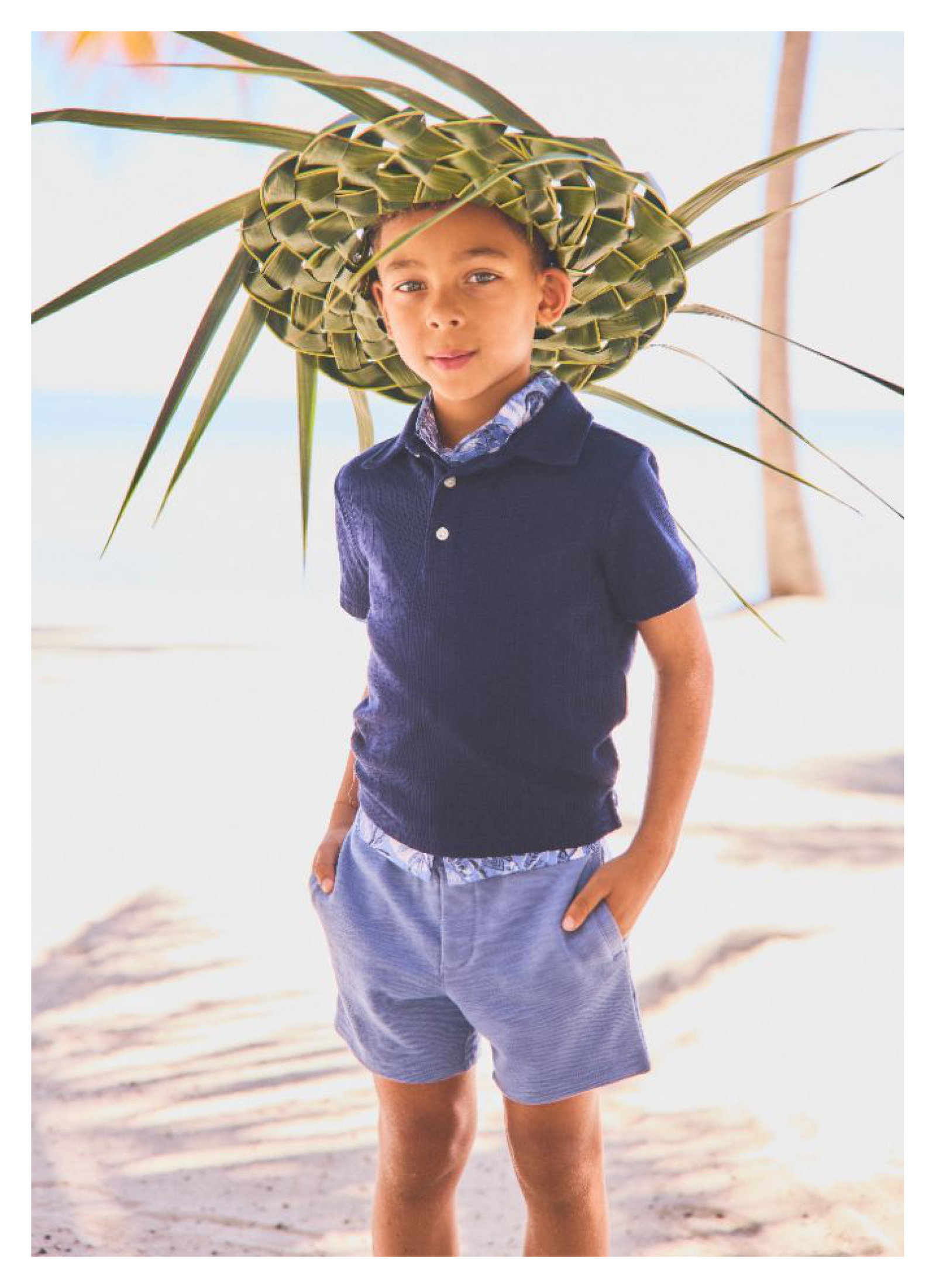 A young boy wearing a woven palm leaf hat, navy polo shirt, and blue shorts standing on a beach