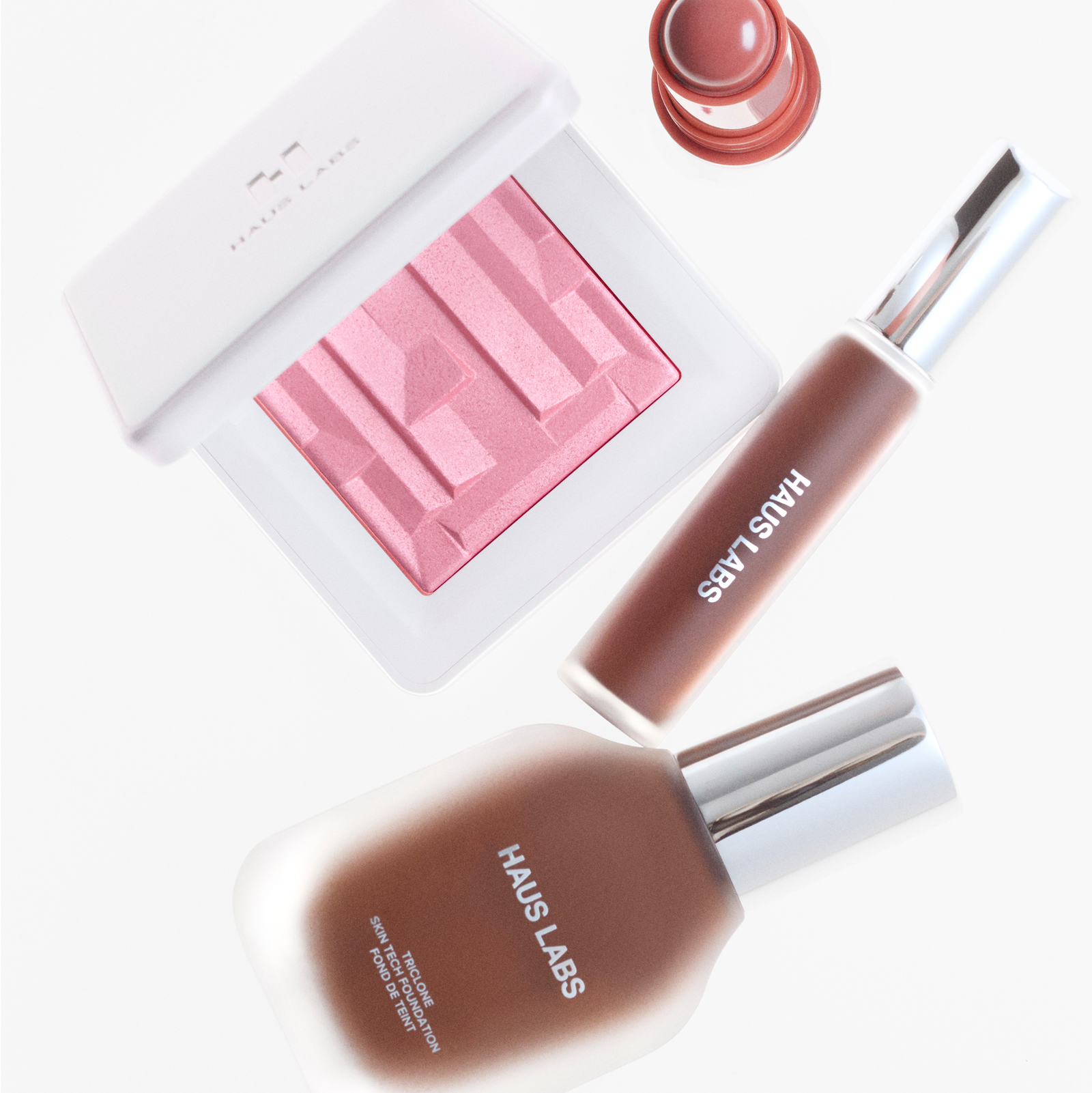 Flat lay of Haus Labs Triclone Skin Tech Foundation and Concealer, Bio-Radiant Gel-Powder Highlighter, and Glassy Blush Balm on a white background.