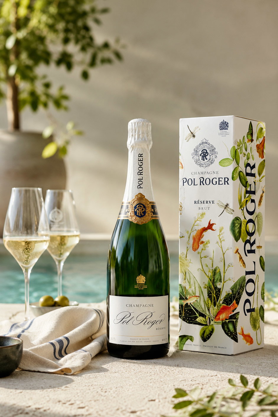 Bottle of Pol Roger Champagne with decorative box and two filled flutes on a sunlit poolside table, summer celebration scene