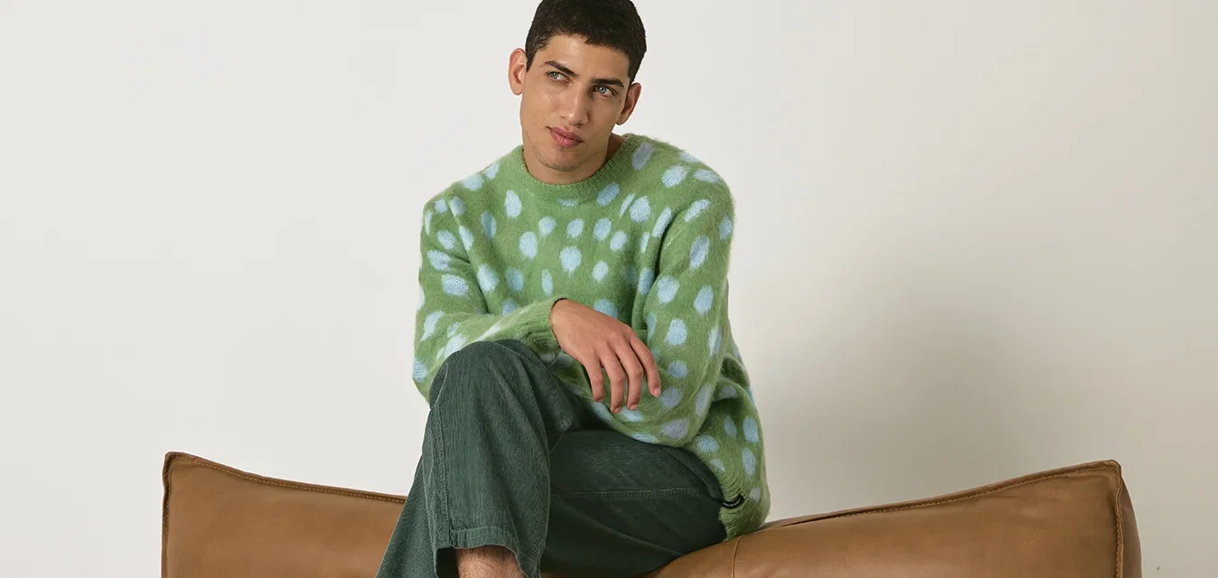Groen en blauwe gepofte trui, groene corduroy broek, wit en grijze sneakers met groene strepen, zittend op een bruin leren bank.