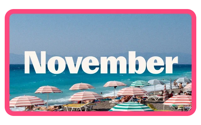 Pebble beach lined with striped beach umbrellas in shades of pink, white, and turquoise overlooking a vivid turquoise-blue sea. Sun loungers and beachgoers are scattered along the shoreline, with distant mountains visible on the horizon under a clear sky. Large white text reading 'November' is prominently displayed across the centre of the image, which is bordered by a bright pink frame.