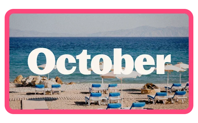 Pebble beach overlooking a clear blue sea, with rows of blue sun loungers and white beach umbrellas arranged along the shoreline. Rocky outcrops sit near the water’s edge, and hazy mountains are visible in the distance across the water. Large white text reading 'October' is centered over the image, which is framed by a bright pink border.