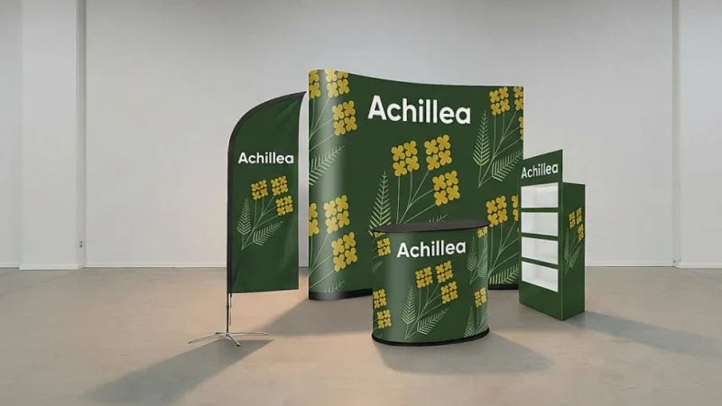 Stand fieristico con sfondo verde, motivi floreali gialli e la scritta “Achillea” su diversi elementi
