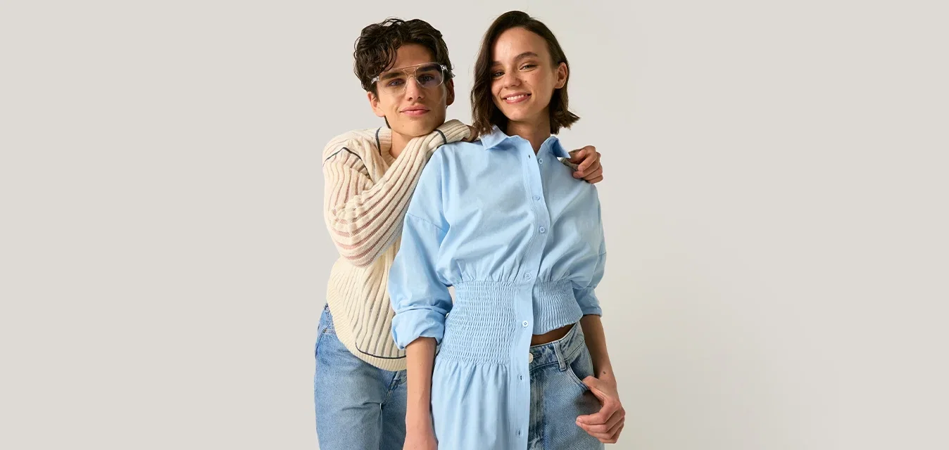 Twee jonge vrouwen staan dicht bij elkaar, één met een bril en een crèmekleurige trui omhelst een ander in een blauwe overhemdjurk, beiden glimlachen tegen een effen achtergrond.