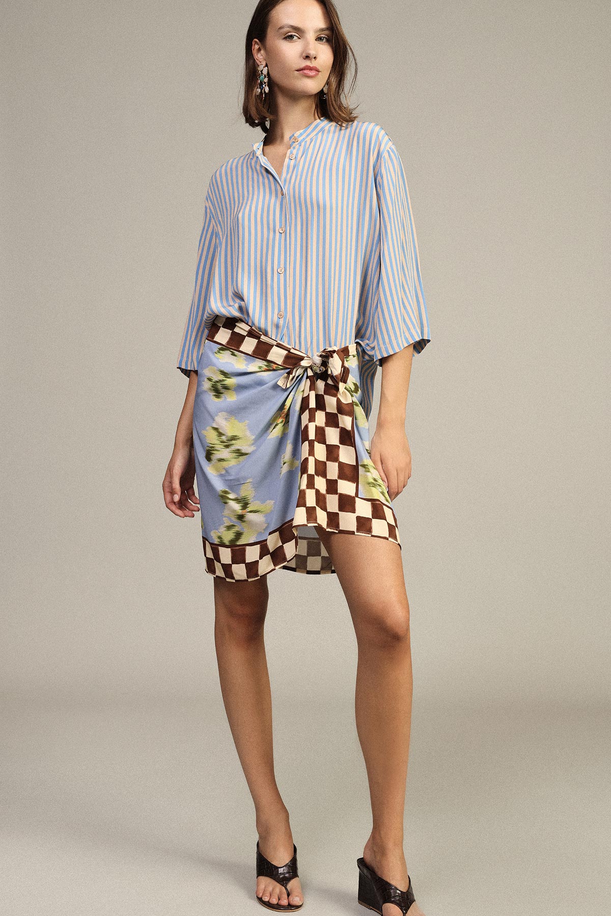 Woman wearing blue striped shirt and floral-checkered wrap skirt tied at the waist, black heeled mules on a neutral studio background