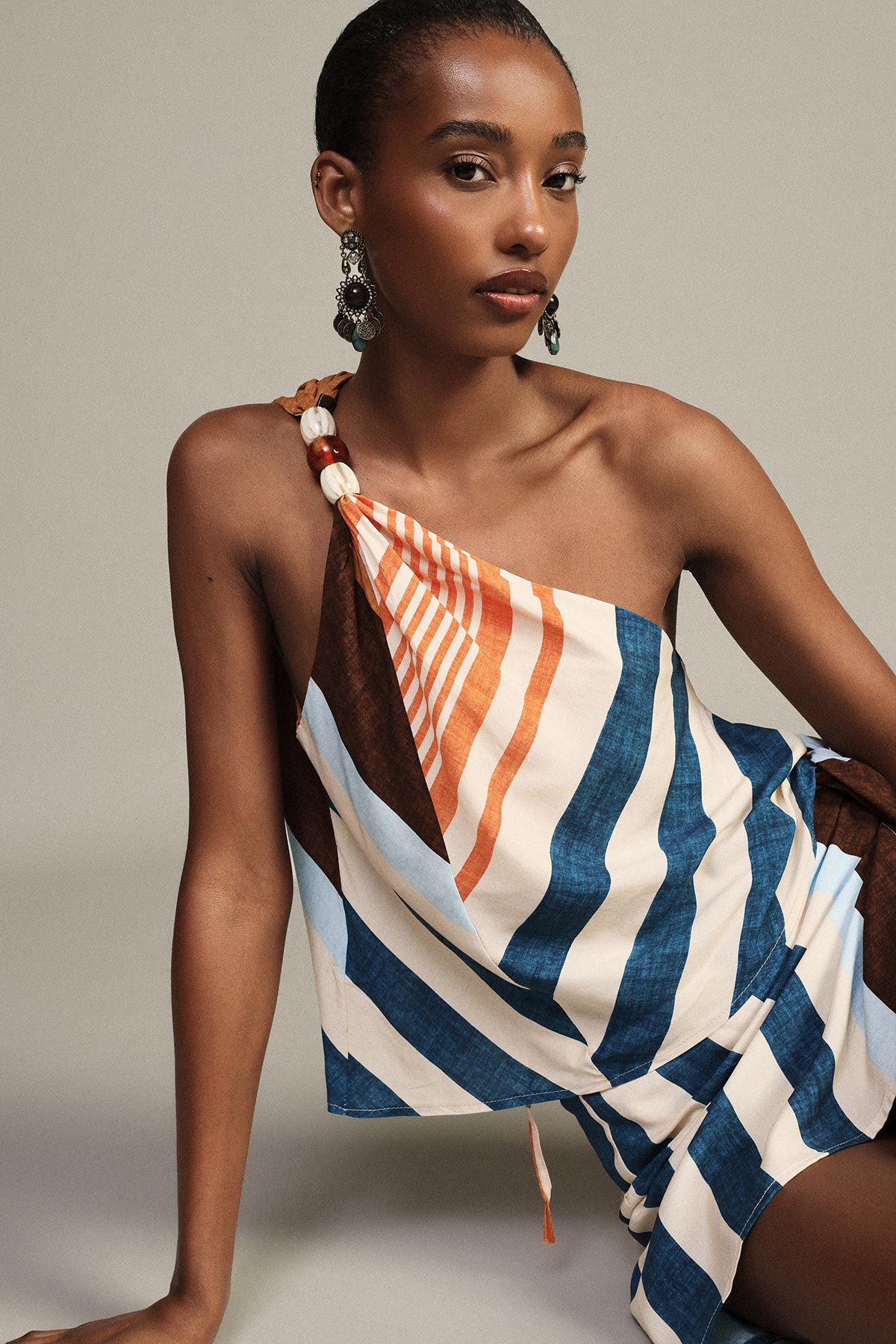 Seated woman model wearing a one-shoulder striped blue, white and orange summer dress with statement drop earrings