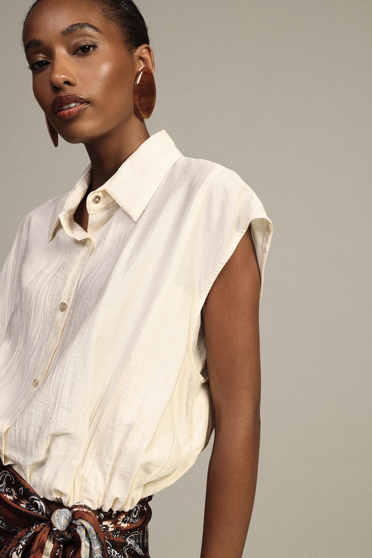 Model modelling sleeveless cream blouse, oversized tortoiseshell hoop earrings and patterned scarf belt on neutral background