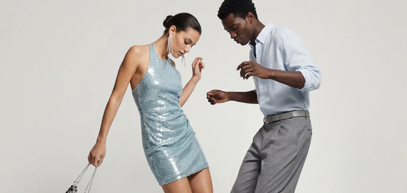 Vrouw in een blauwe paillettenjurk en man in een overhemd en nette broek dansen samen, beiden glimlachend en in beweging.
