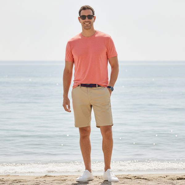 Man wearing coral T-shirt, khaki shorts and sunglasses standing on a sandy beach by calm sea, casual men's summer outfit