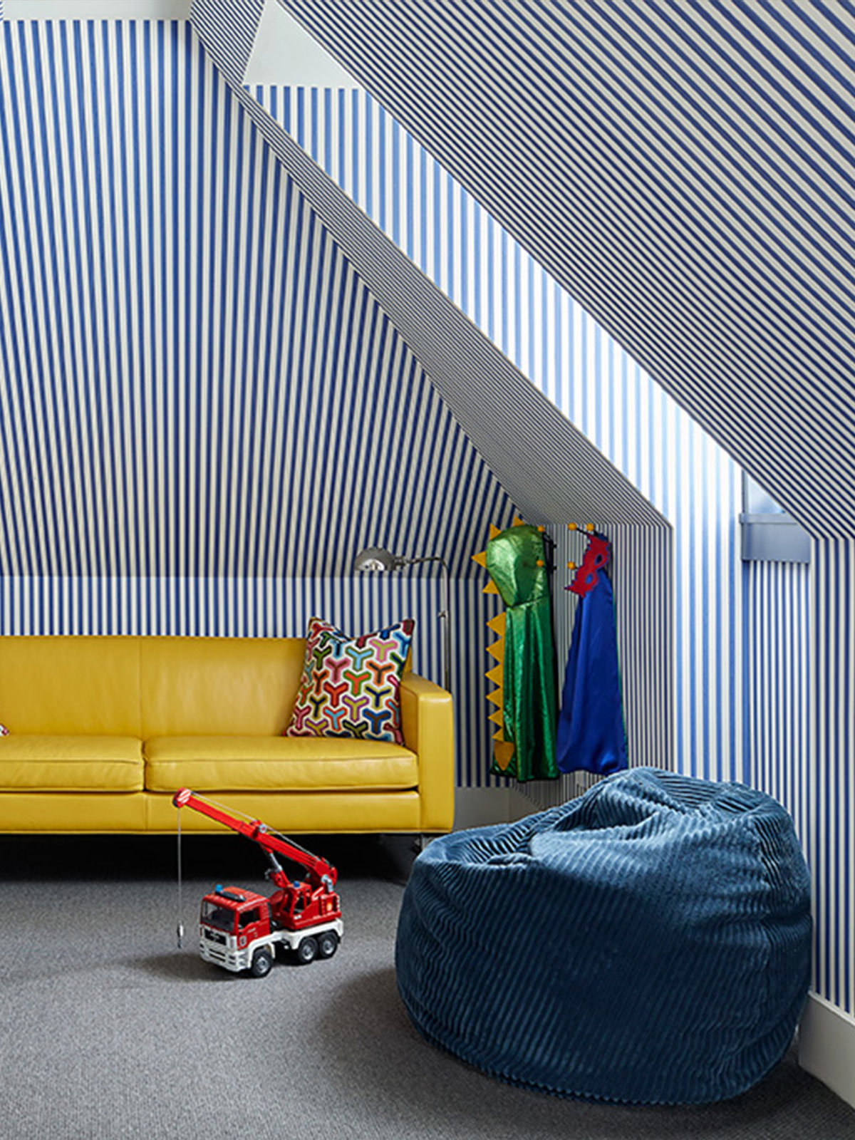 A playful attic playroom wrapped in blue and white striped wallpaper, with a bright yellow sofa, toys and beanbag creating a cheerful space for children.