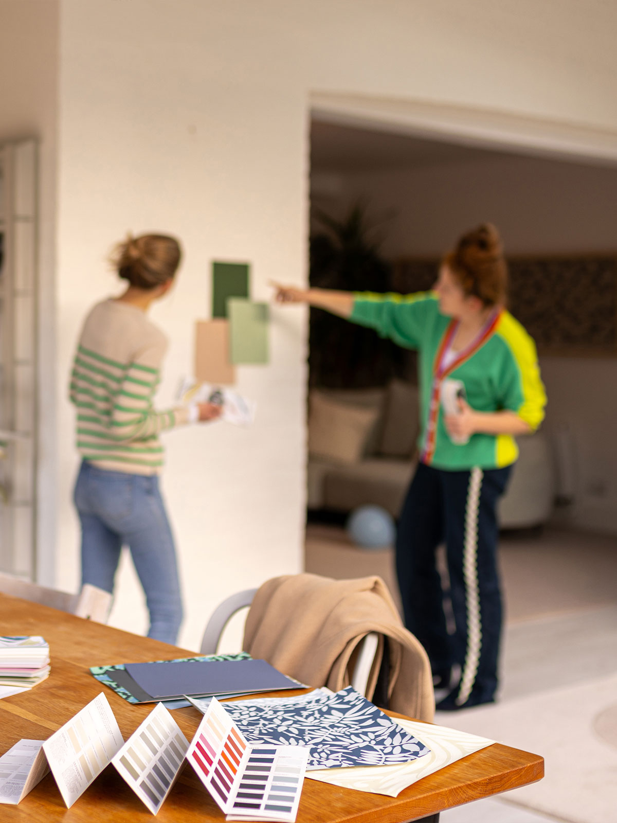 Two people discuss paint colours in a bright room, with sample cards and colour charts on a wooden table.