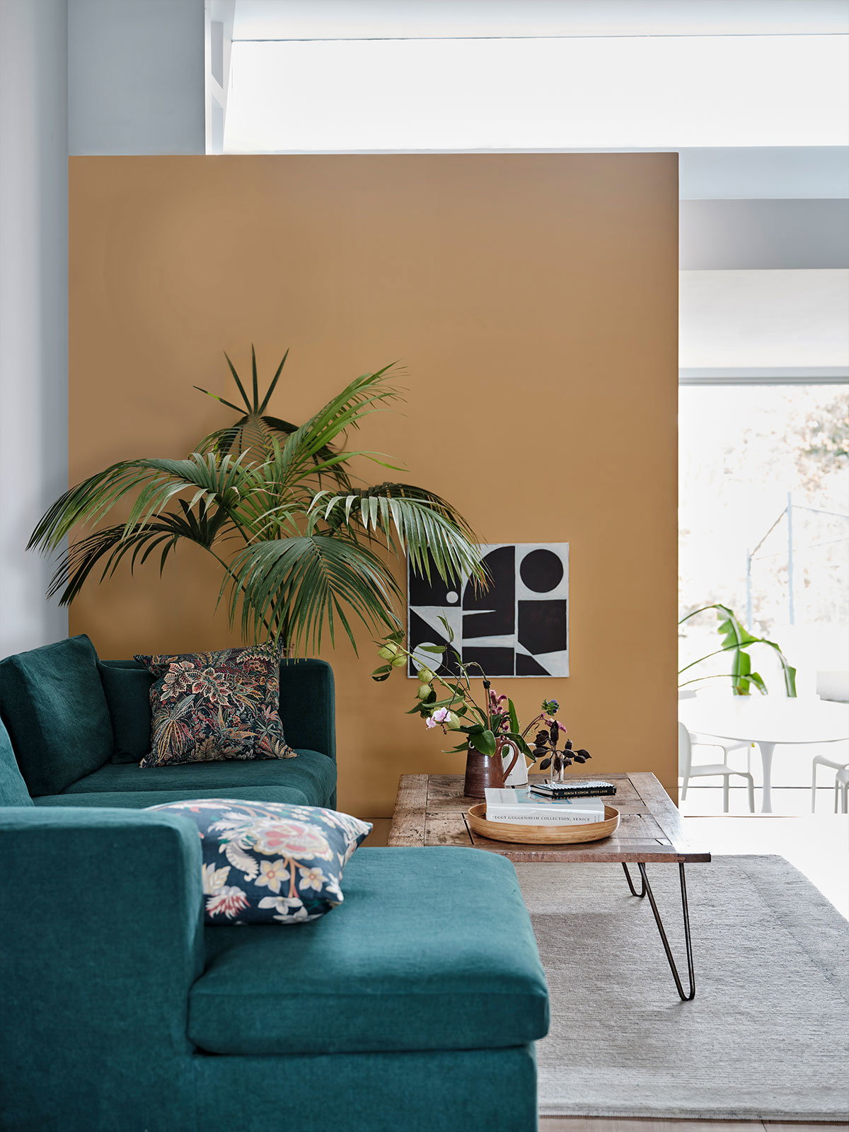 A modern living room with a teal sofa, mustard feature wall, large palm plant, and wooden coffee table with books and flowers.