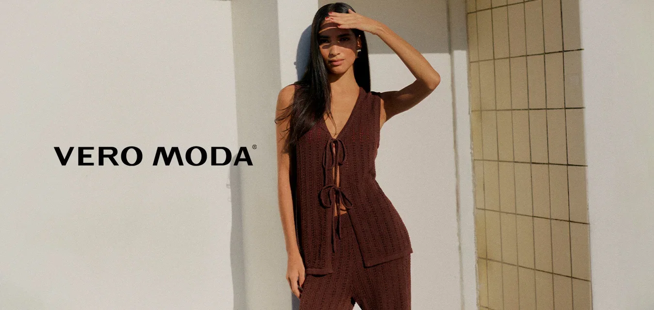 Donna con top marrone lavorato a maglia senza maniche e pantaloni in piedi scalza su un patio soleggiato, protegge gli occhi con la mano contro il cielo azzurro chiaro.