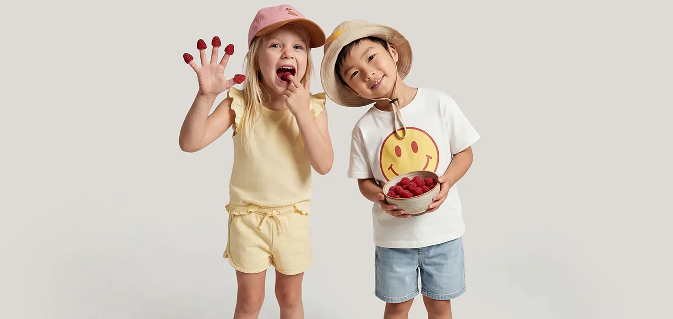 Twee kinderen staan tegen een effen achtergrond; de een draagt gele korte broek en een roze hoed, de ander draagt blauwe korte broek, een wit shirt en een beige hoed en houdt een speelgoed vast.