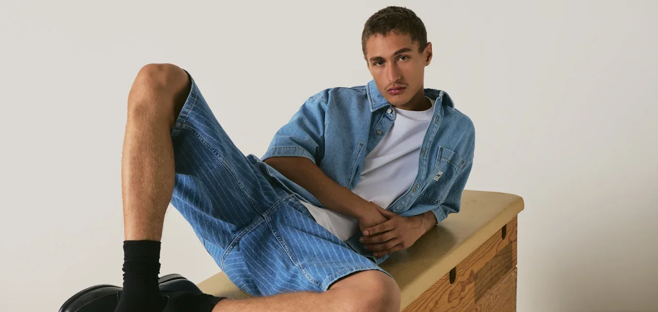 Jonge man die achterover leunt op een houten turndoos, gekleed in denim shorts, denim overhemd, wit t-shirt, zwarte sokken en zwarte loafers.
