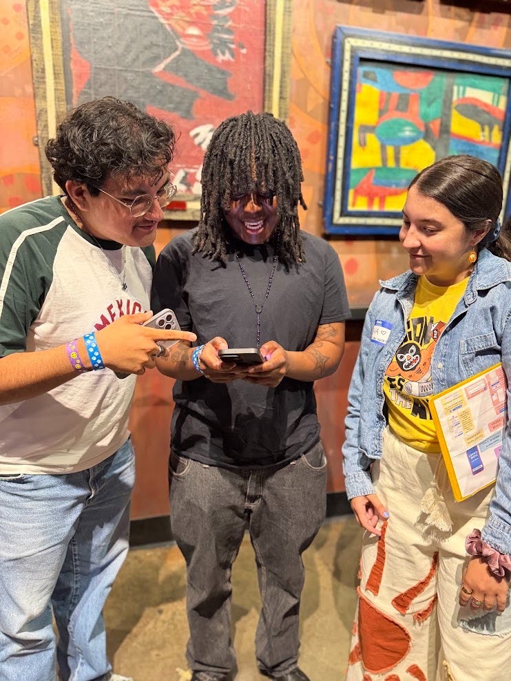Three people standing side by side, all smiling and looking at a phone that the person in the middle is holding