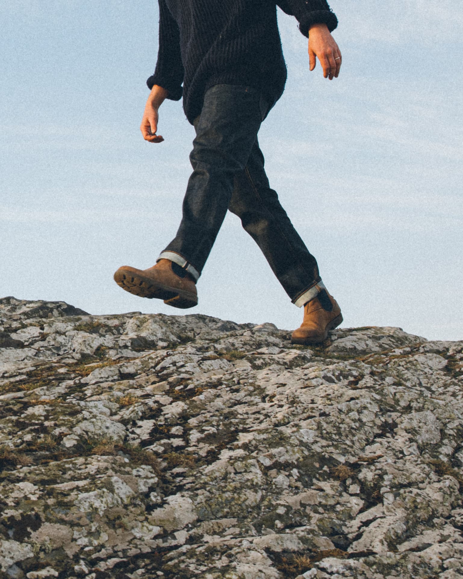 Shoulder down photo of man in navy wool jumper, selvedge jeans, and brown boots taking a step along rocks. Blue sky.