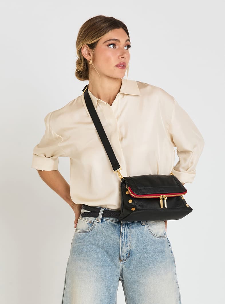 Model wearing the Hammitt Beverly Med in Black/Brushed Gold Red Zip pebbled leather as a crossbody, featuring signature rivets, brushed gold hardware, and the iconic red zipper detail.