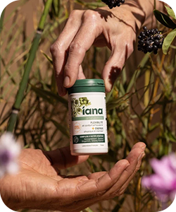 Two hands passing a small green-lidded Iana supplement bottle amongst wildflowers and grasses