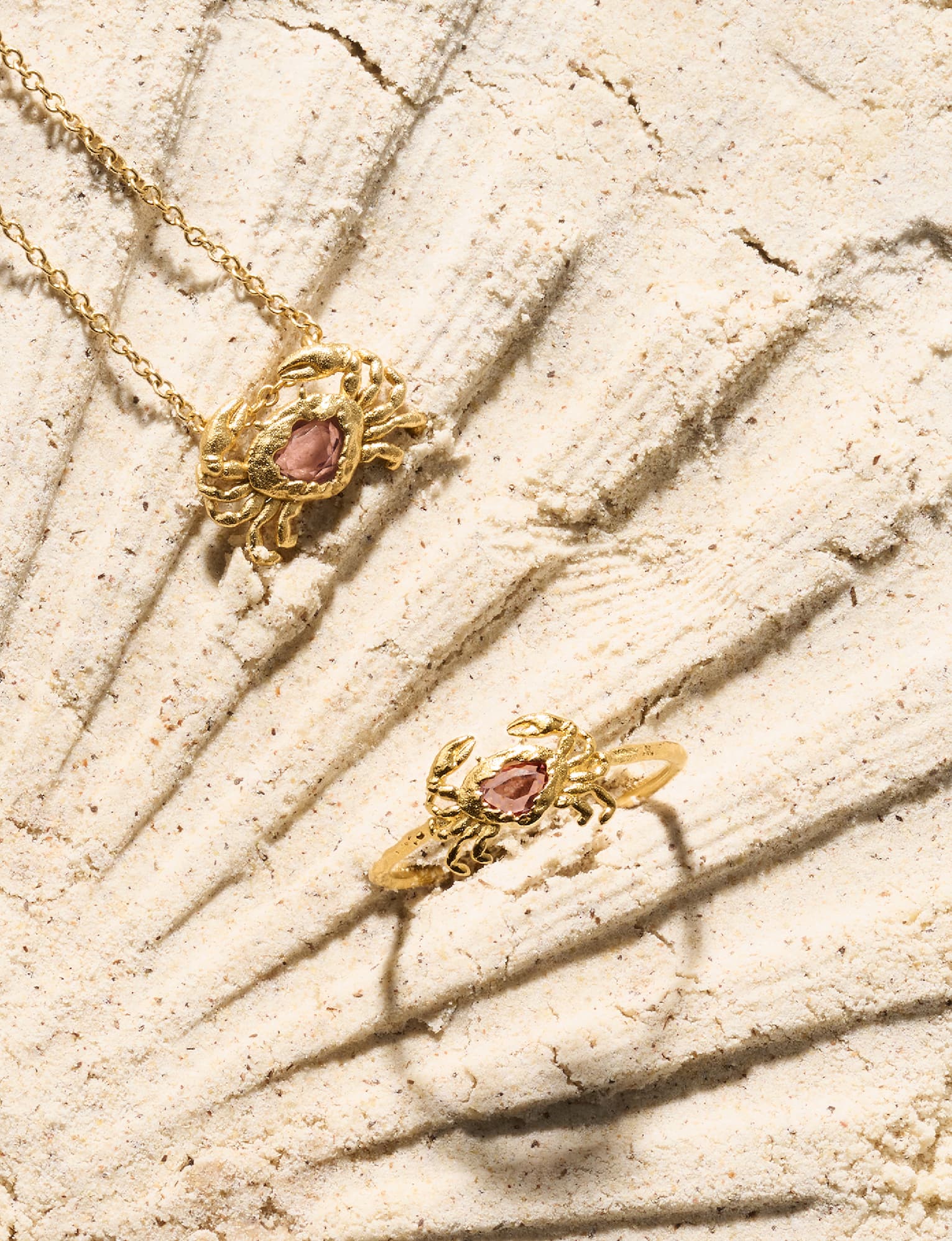 Gold crab pendant necklace and matching crab ring, each set with a pink stone, resting on sand