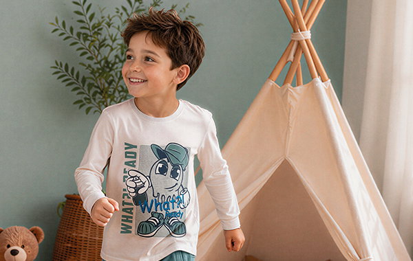 Junge trägt Jungen-Langarmshirt, weiß mit blauen Motiv Spielzimmer mit Holzfiguren im Hintergrund.