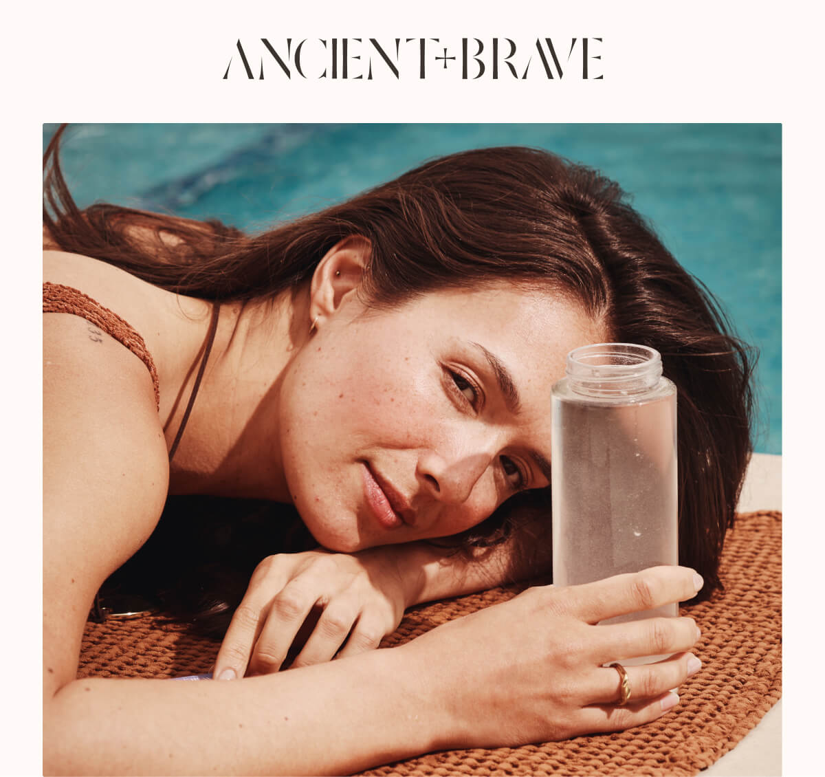 Woman lying poolside on a woven towel, resting her head on her arm while holding a bottle of Ancient + Brave collagen water.