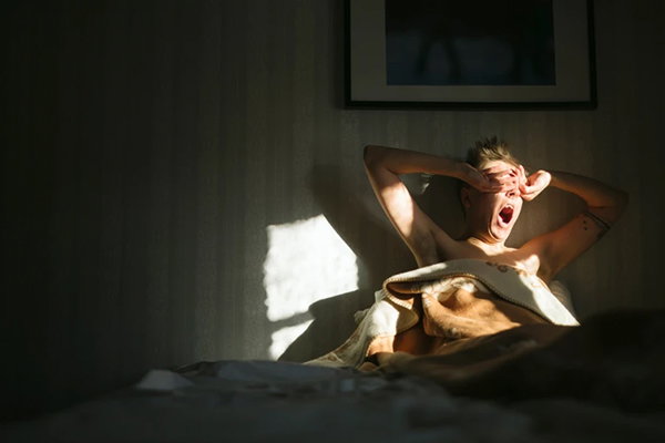 A person sitting up in bed, yawning, stretching and covering their eyes as bright morning sunlight hits their face in an otherwise darkened room.