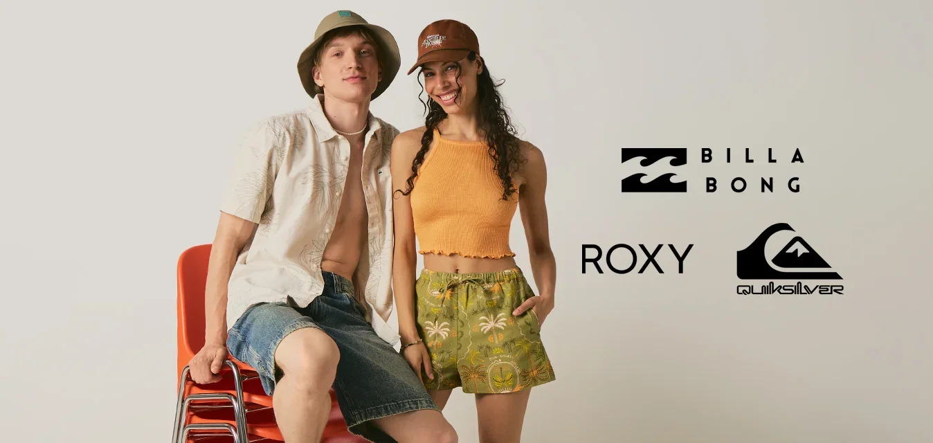 Joven con pantalones cortos y sombrero tipo pescador sentado sobre sillas rojas apiladas junto a una mujer sonriente con top naranja y pantalones cortos con estampado tropical.