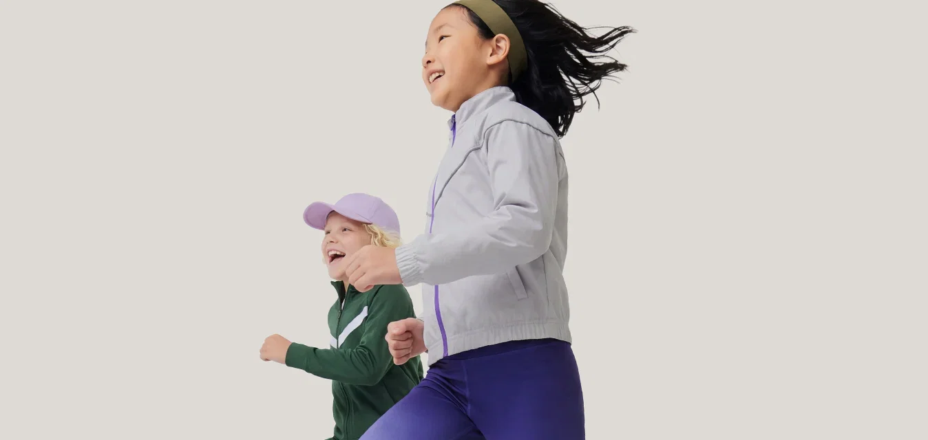 Dos niños corriendo lado a lado, vestidos con ropa deportiva y zapatillas, una niña con chaqueta y leggings y la otra con gorra y chándal.